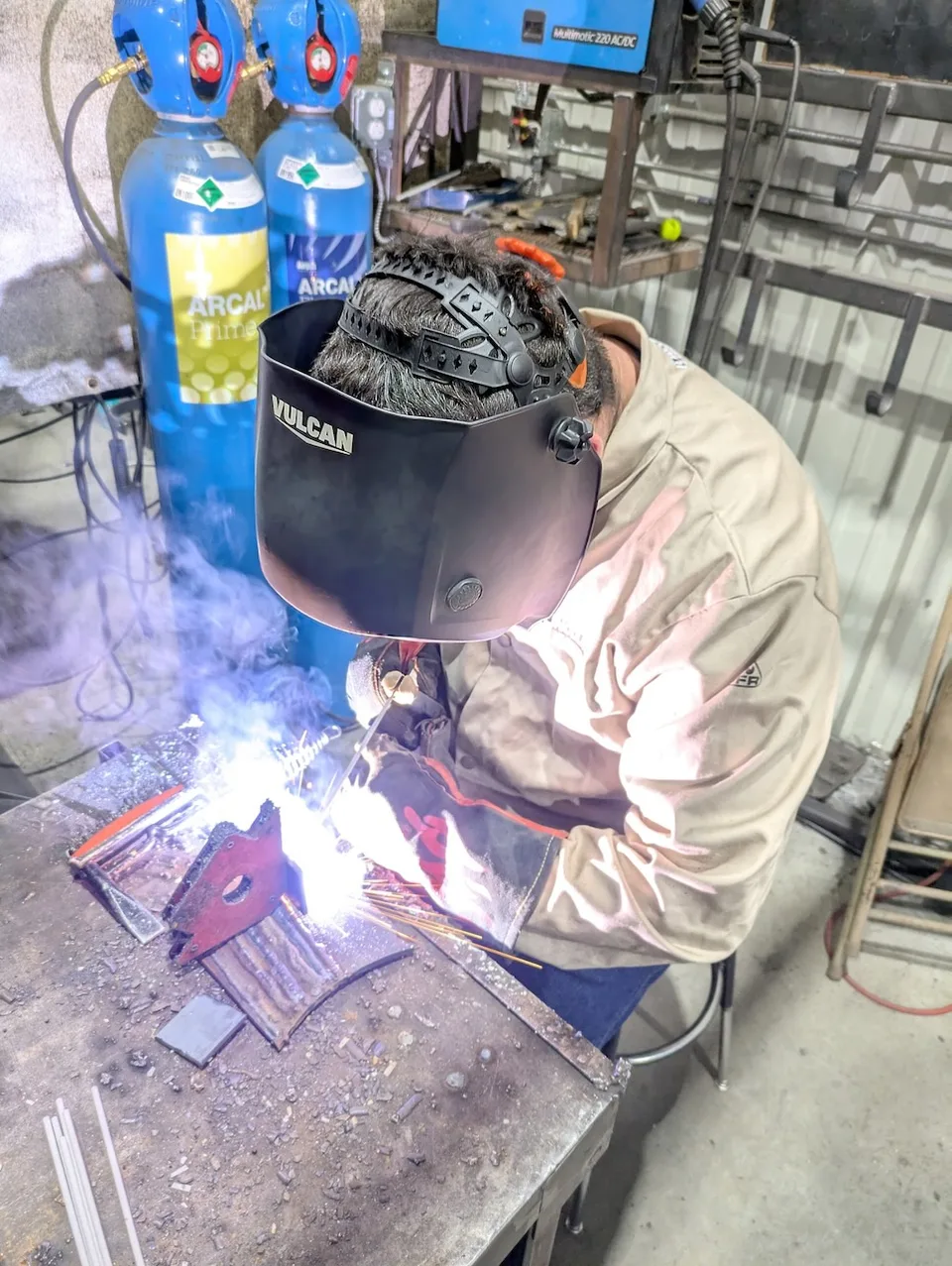 A person wearing a welding helmet and gloves while welding at a workbench.